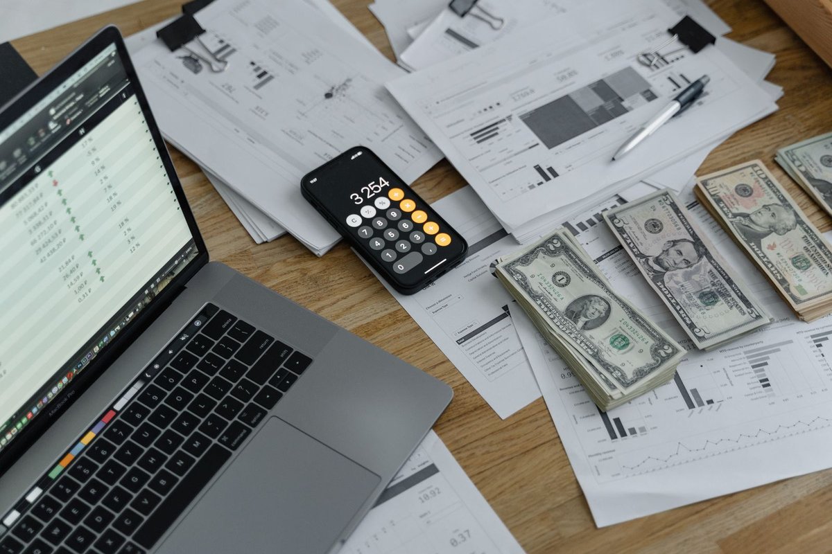 Wooden desk with laptop spreadsheet, phone calculator, banknotes, and financial chart printouts