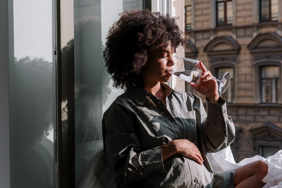 A pregnant woman sitting by a window holding a glass of water